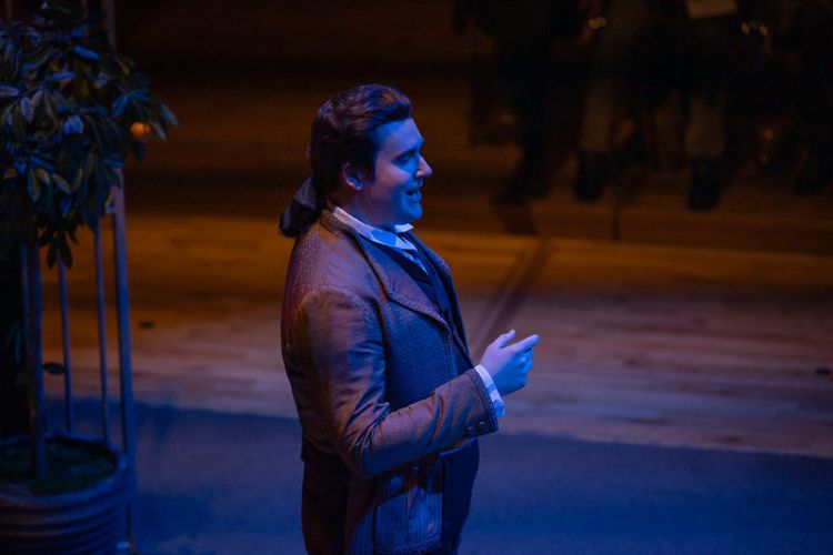 A male opera singer in historical costume and wig, singing on a darkened stage